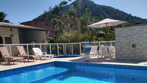 A swimming pool with lounge chairs and an umbrella, surrounded by a stone wall and a mountain in the background. - Canto Verde Aptos III (Caraguatatuba)