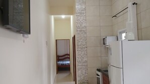 A narrow kitchen with a refrigerator, a microwave, and a small oven. A television is mounted on the wall. The kitchen has white tiled walls and floor. - Canto Verde Aptos III (Caraguatatuba)