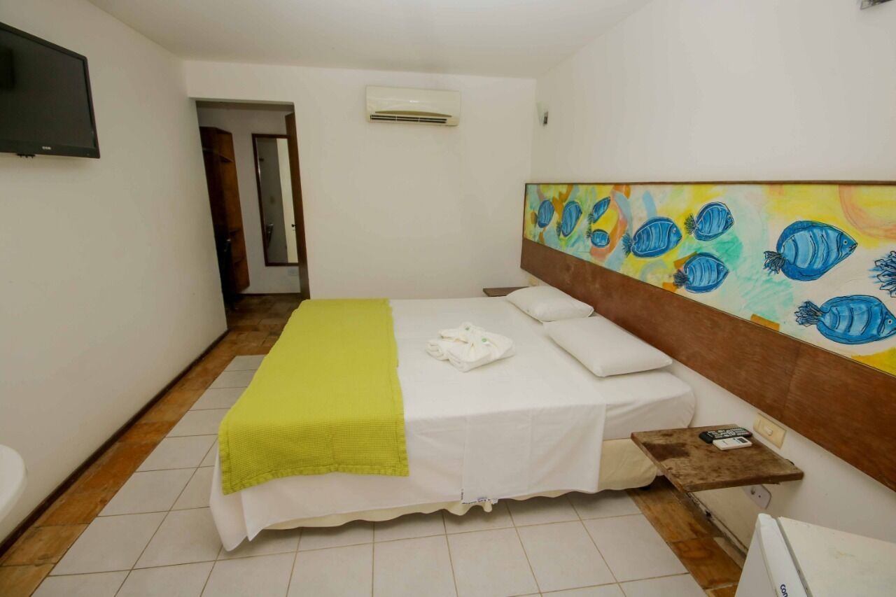A hotel room with a bed, a painting of fish on the wall, a TV, and an air conditioning unit.