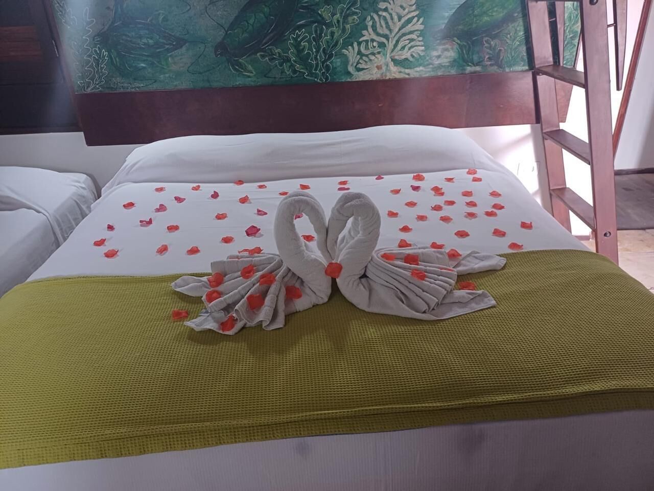 A hotel room with a bed featuring a white bedspread, a green throw blanket, and two towels arranged in a swan shape with red rose petals.