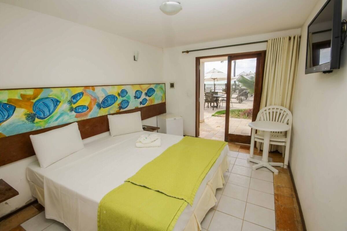 A hotel room with a large bed, a painting on the wall, a TV, and a balcony with outdoor seating.