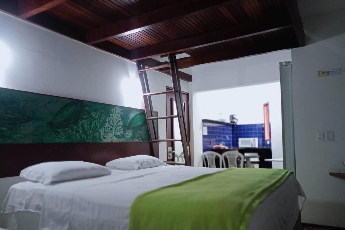 A bedroom with a bed, a green blanket, a wooden ladder, and a view of a kitchen area.