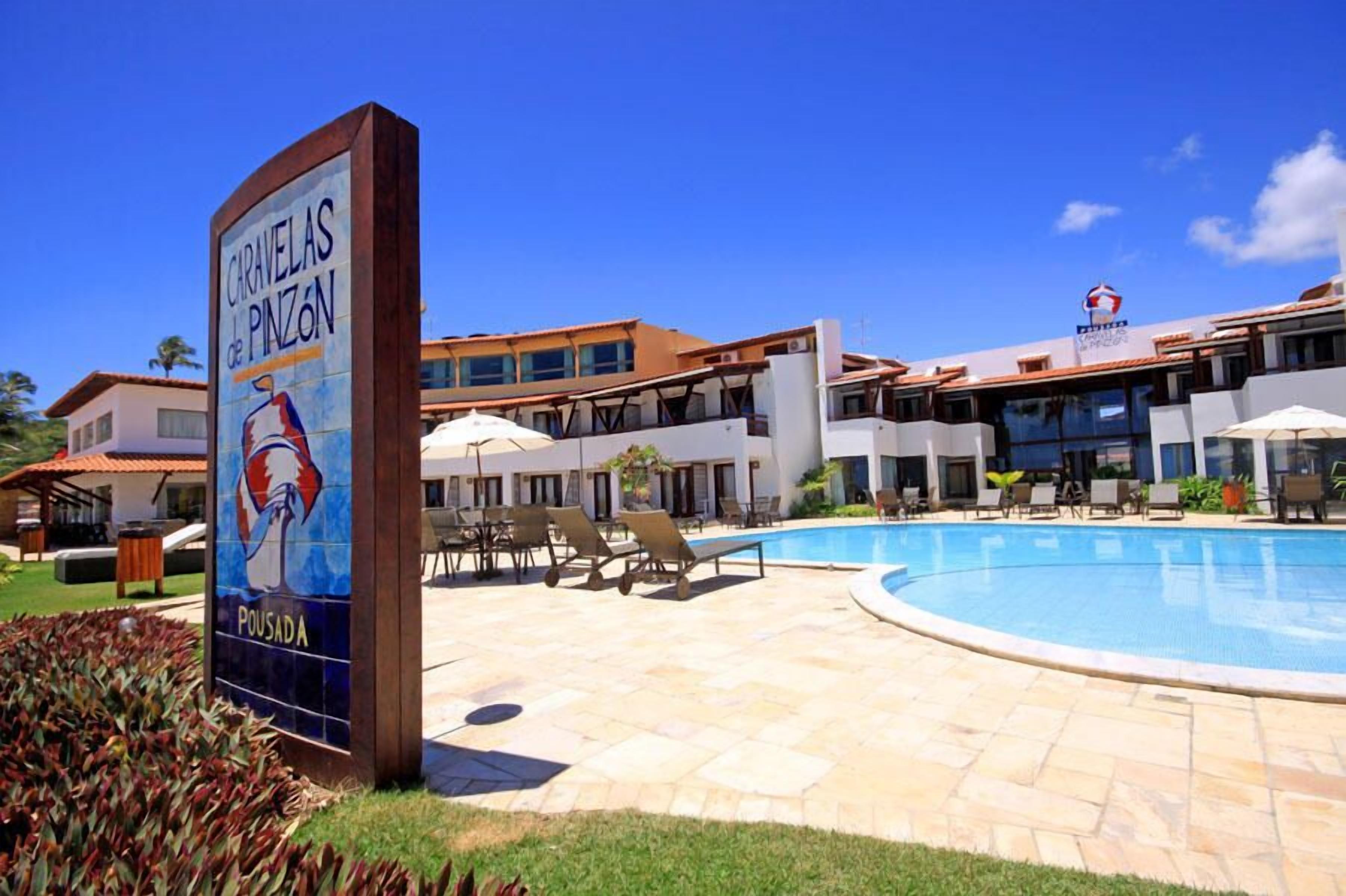 A hotel with a swimming pool, lounge chairs, and a sign for 'Caravelas de Pinzon'.