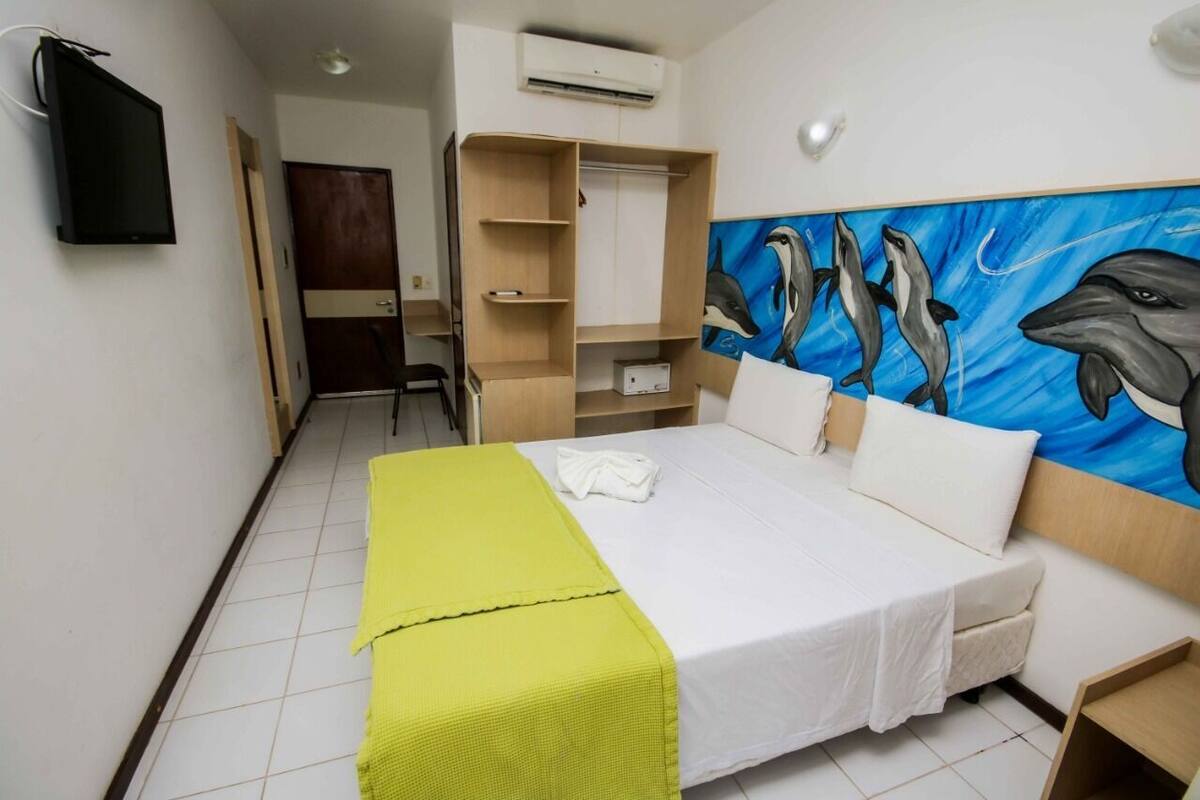 A hotel room with a bed, a painting of dolphins, a wardrobe, and a desk.
