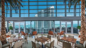 A spacious indoor area with multiple tables and chairs, large windows offering a view of a cityscape and the sea, and palm trees. - 1 Bedroom Brickell Condo (Miami)