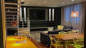 A modern living room with a flat-screen TV, a sofa, a dining table with chairs, and a staircase with bookshelves.