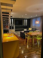Un salon moderne doté d’un téléviseur à écran plat, d’un canapé, d’une table à manger avec des chaises et d’un escalier avec des étagères.