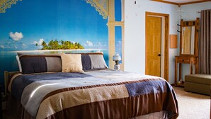 A bedroom with a large bed, a ceiling fan, a wooden door, and a painting of a tropical beach scene. - Shane Acres Country Inn (Juneau)