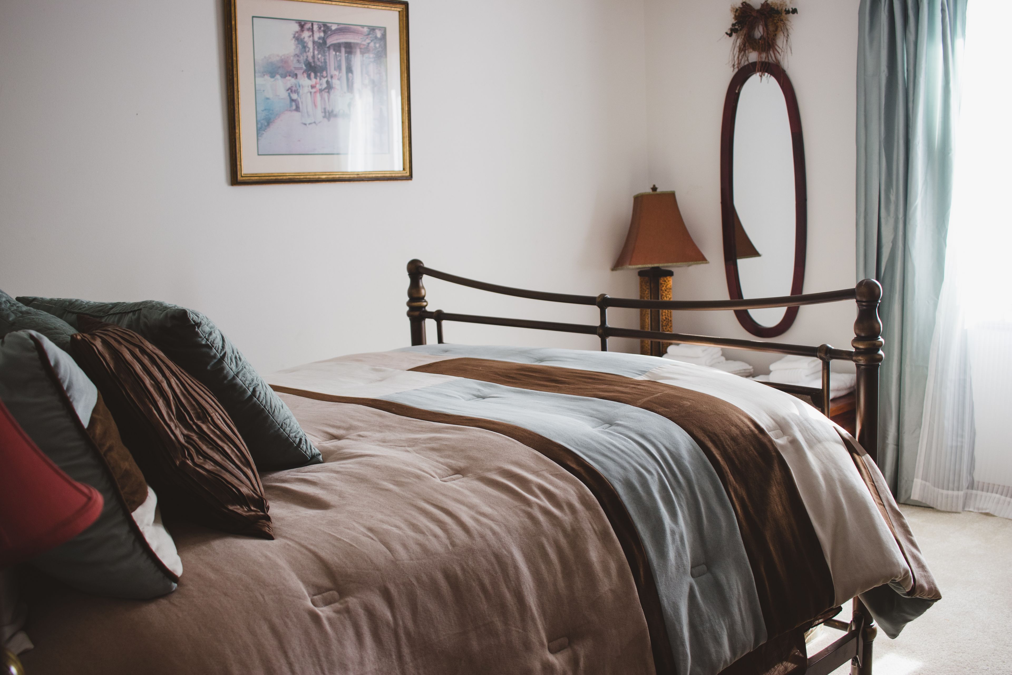 Una cama bien hecha con estructura de madera, una lámpara de noche, un cuadro en la pared y una ventana con cortinas.