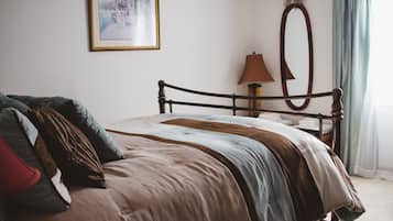 Una cama bien hecha con estructura de madera, una lámpara de noche, un cuadro en la pared y una ventana con cortinas.