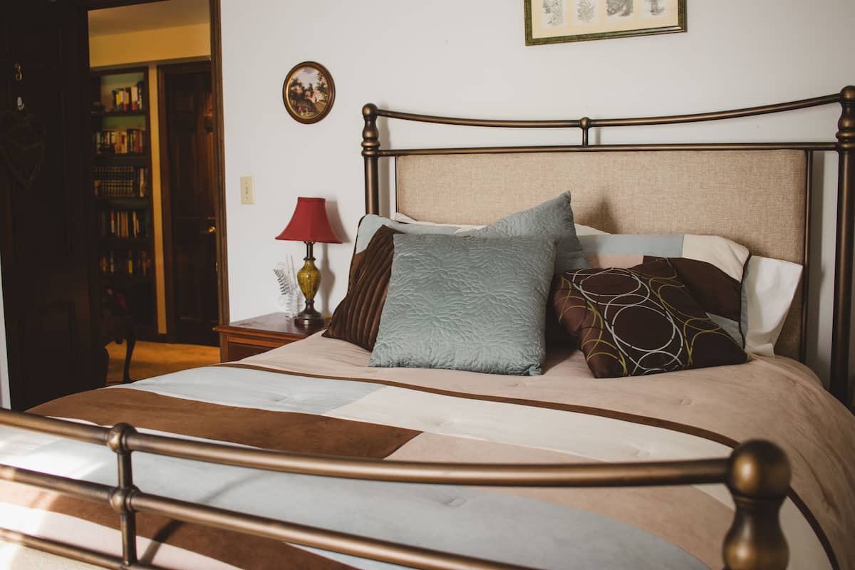 Una cama bien hecha con cabecera de madera, una mesita de noche con lámpara y una estantería al fondo.