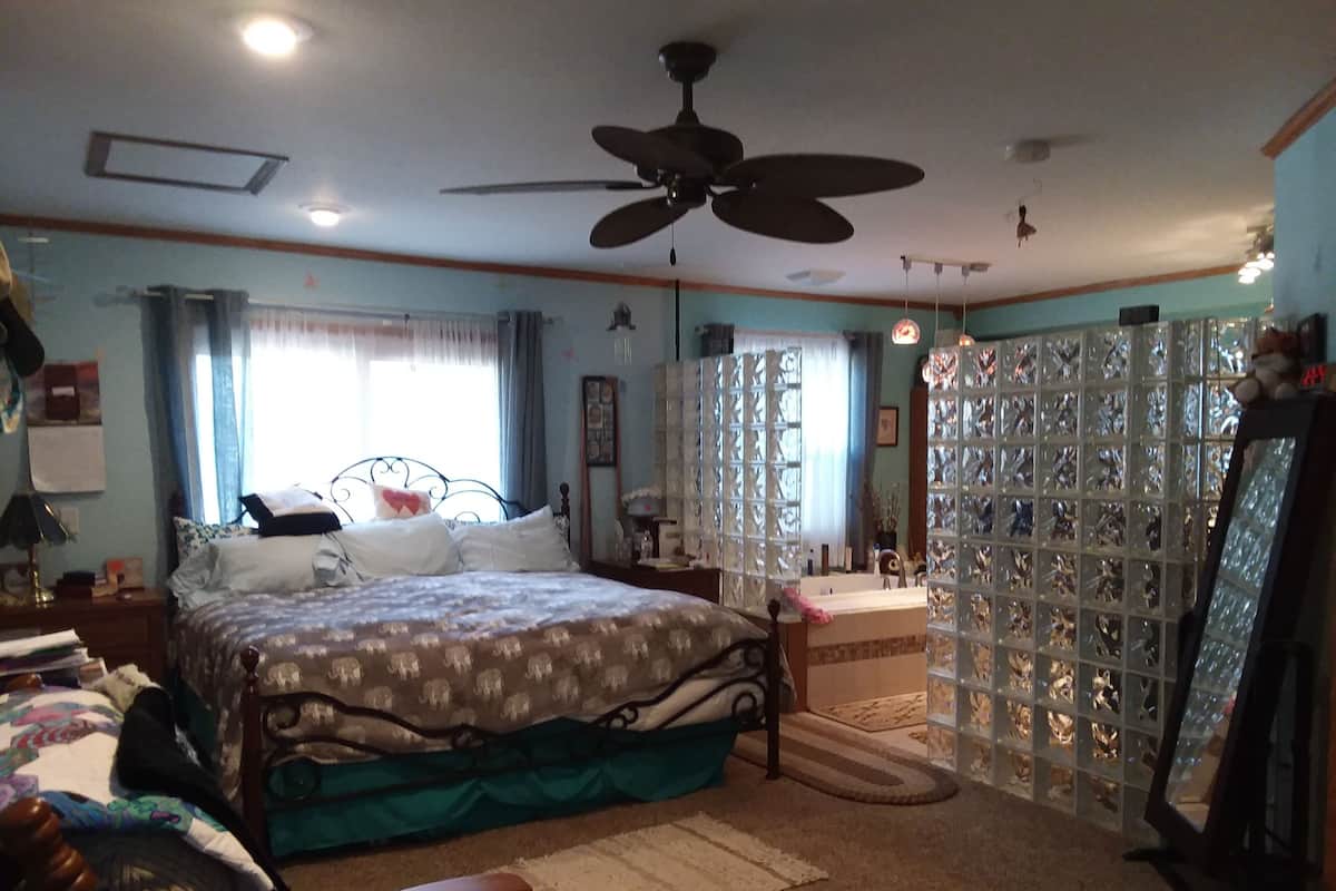 Un dormitorio con cama, ventilador de techo, ventana con cortinas, espejo y radiador.