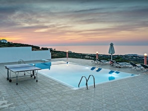 A rooftop pool with a table tennis table, lounge chairs, and a view of the sea at sunset.
