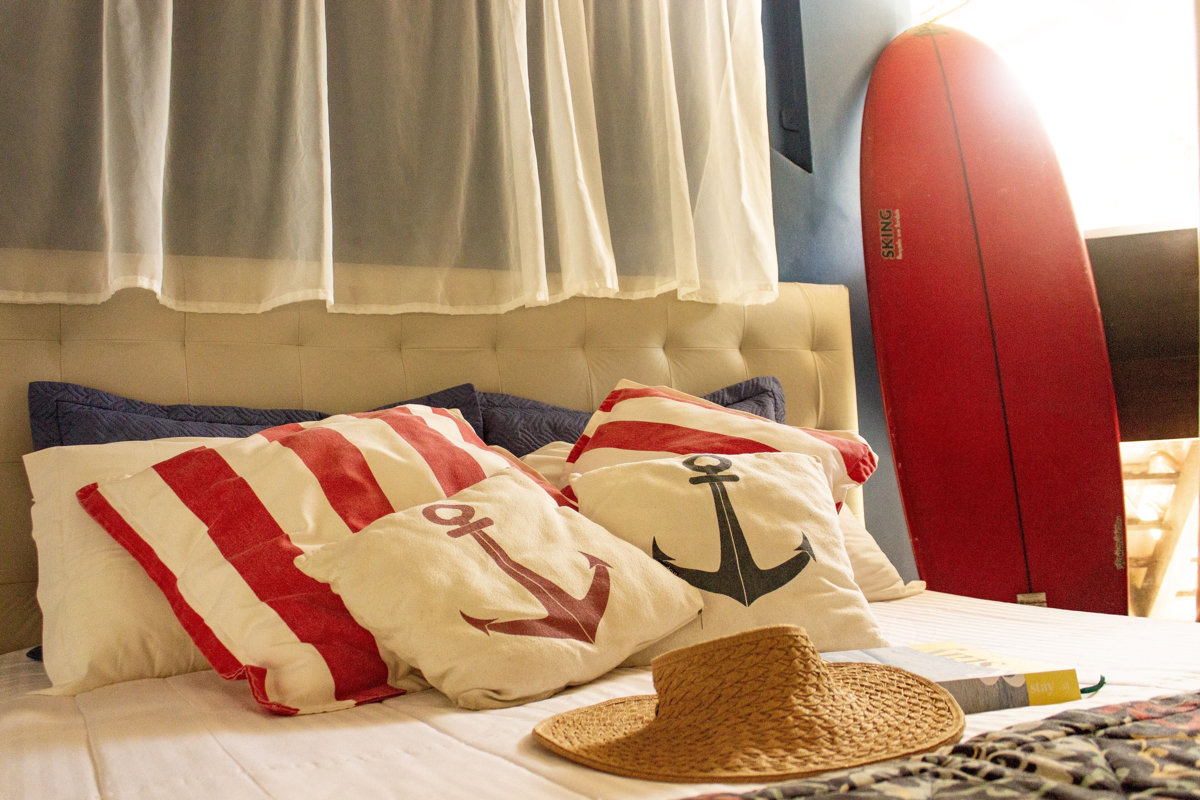 Ein Schlafzimmer mit einem Bett, Kissen mit Anker-Motiven, einem Strohhut und einem roten Surfbrett.