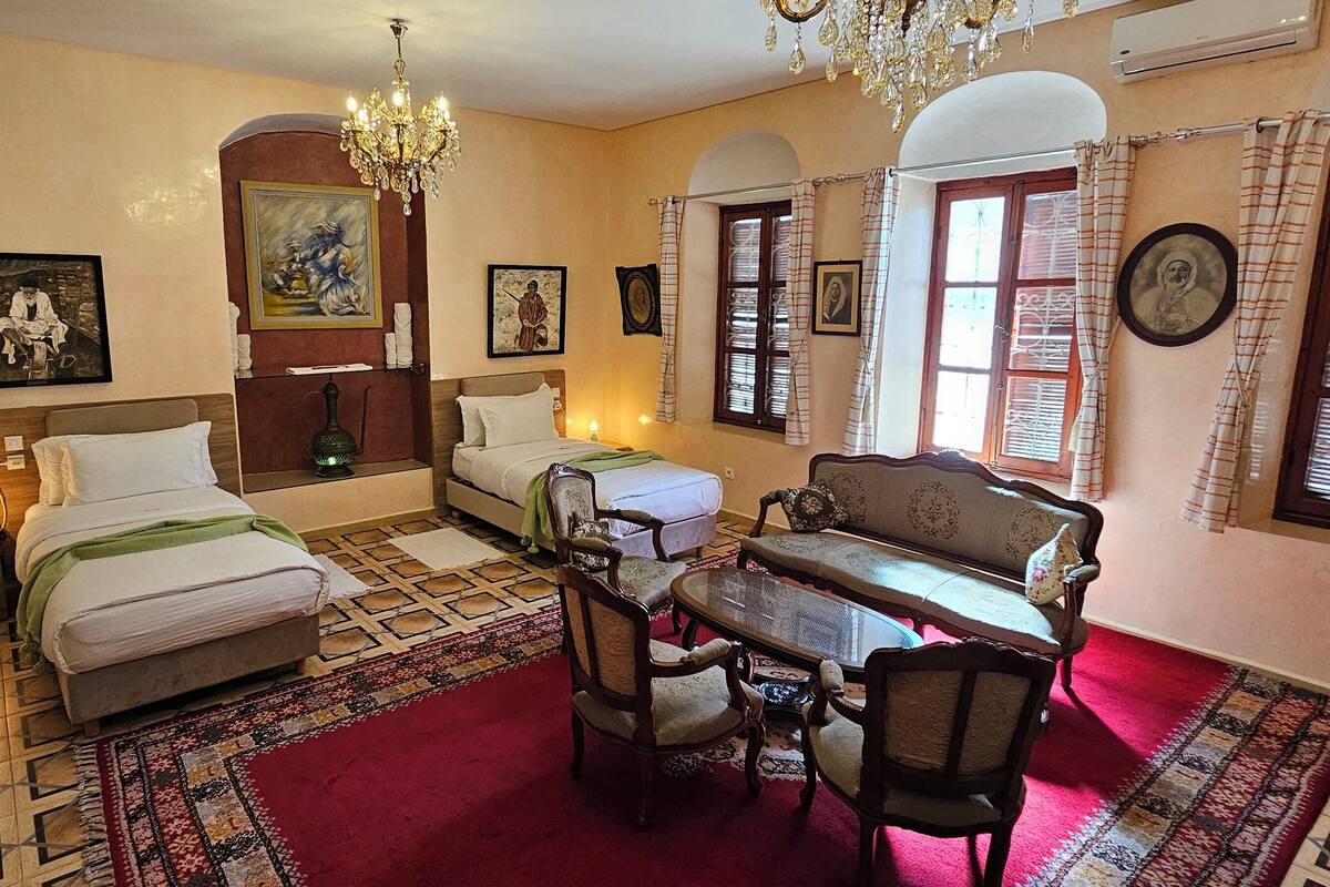 Habitación de hotel con dos camas, una mesa de comedor con sillas, una araña y obras de arte en las paredes.