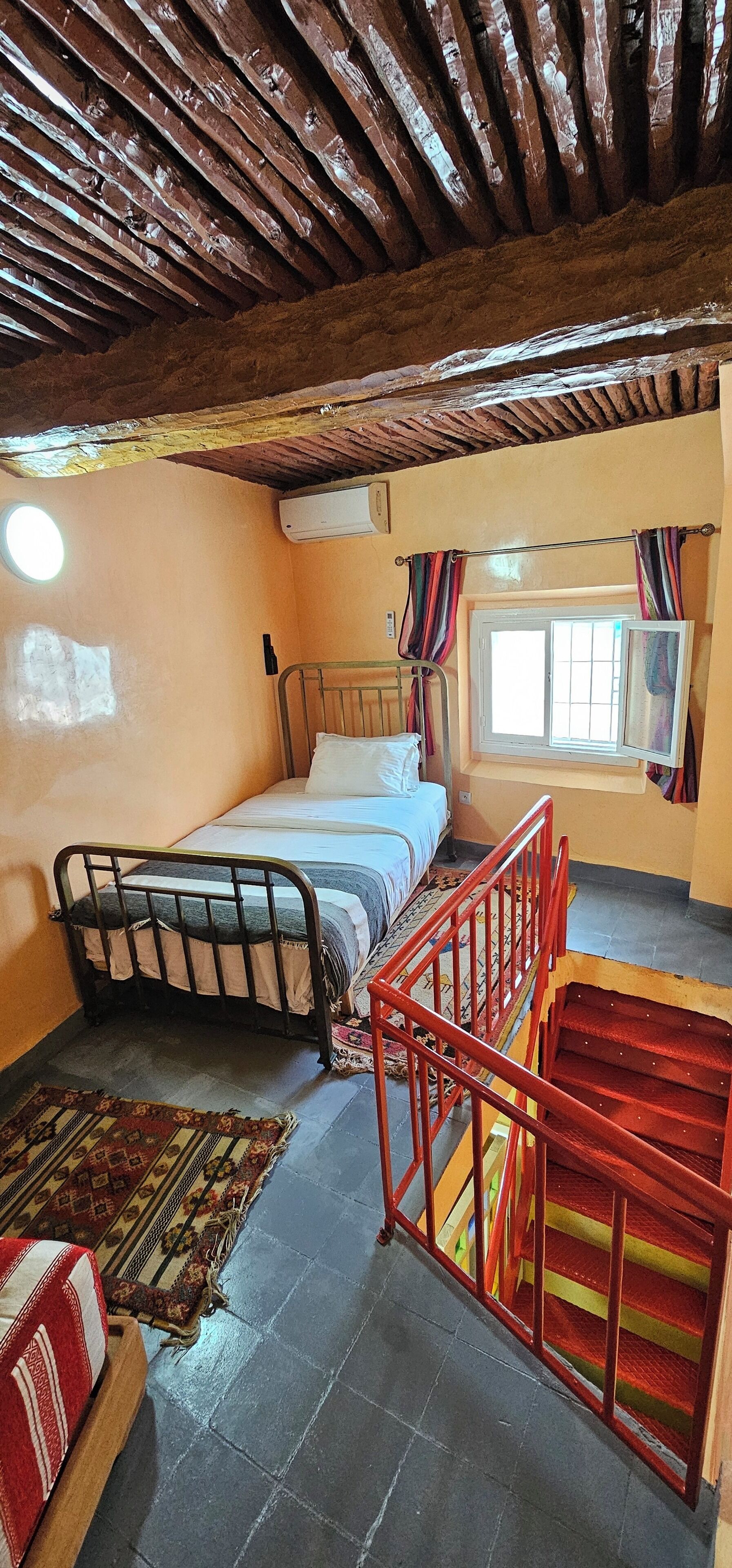 Ein Schlafzimmer mit Bett, einem Fenster mit Vorhängen, einer Treppe und einem kleinen Teppich.