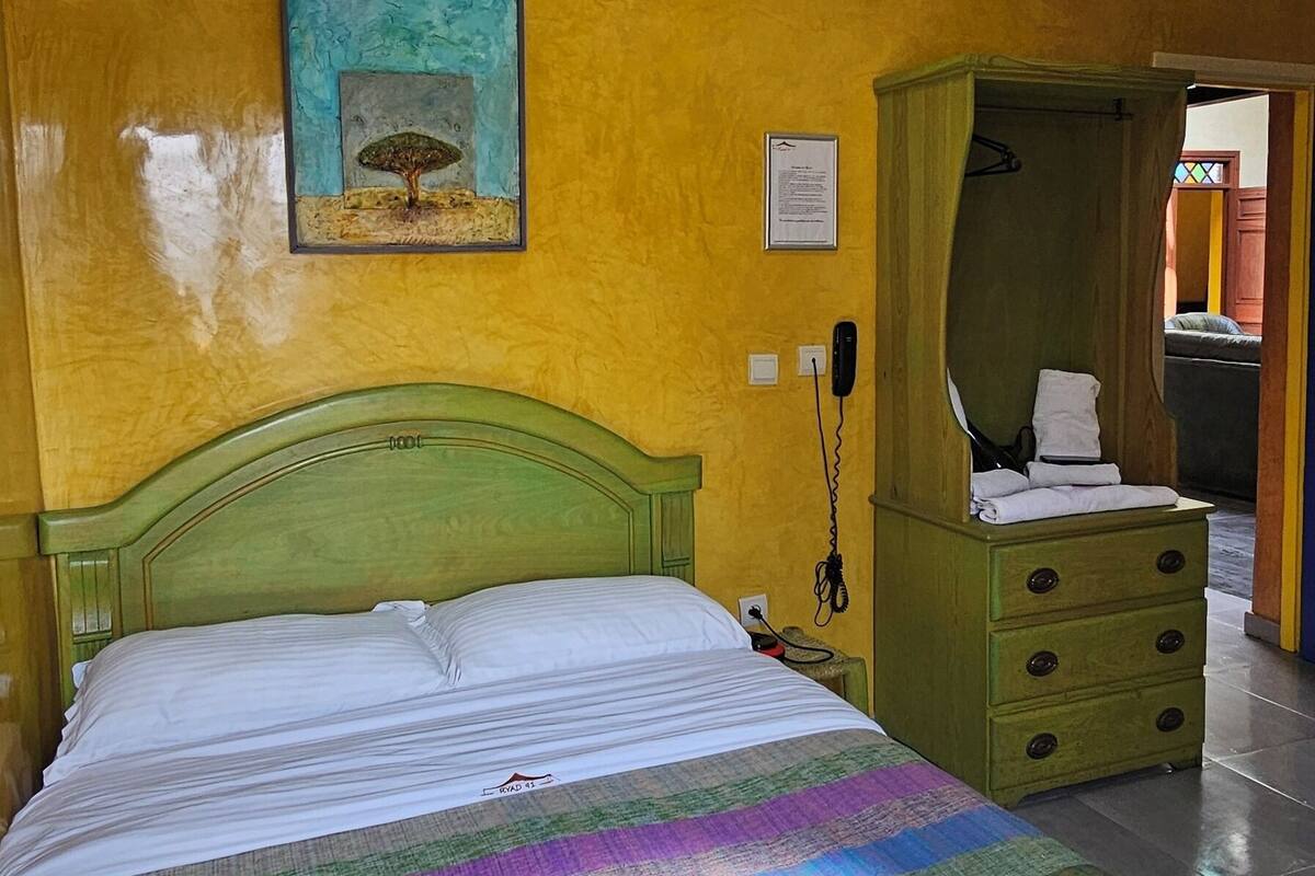 Una habitación con una cama, un aparador verde, un cuadro y una puerta que da a otra estancia.