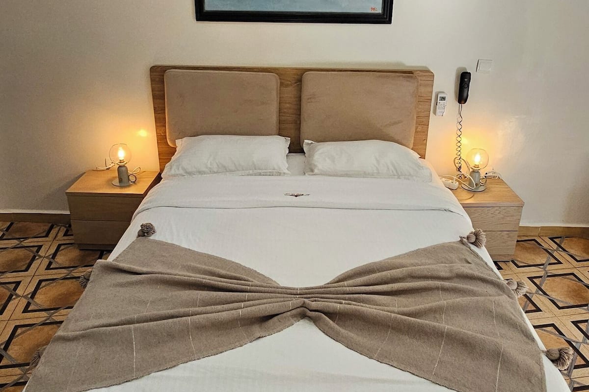 Una cama bien hecha con cabecera, un cuadro enmarcado en la pared, lámparas de noche y una alfombra estampada en el suelo.