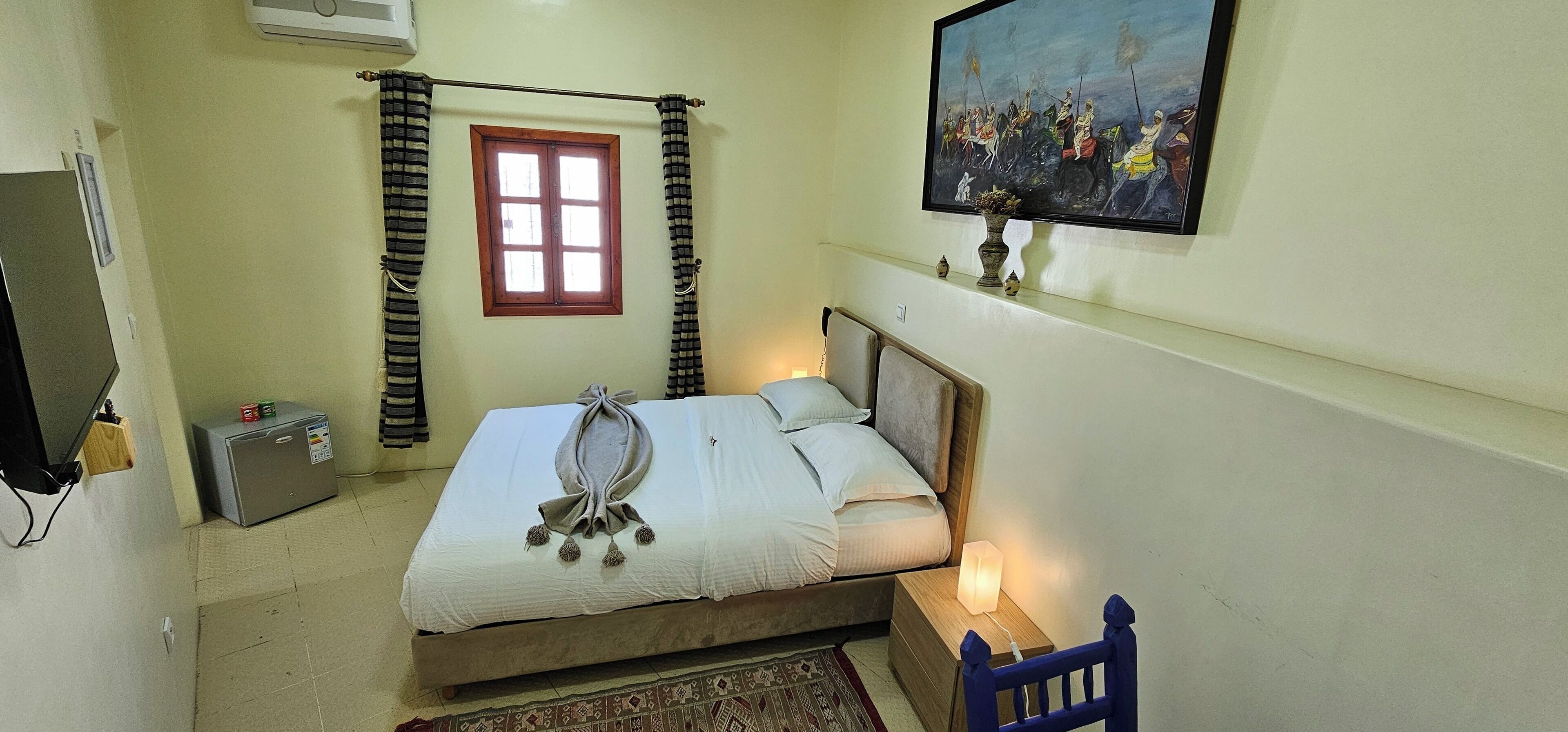 A bedroom with a bed, a nightstand with a lamp, a window with curtains, a radiator, a small heater, and a painting on the wall.