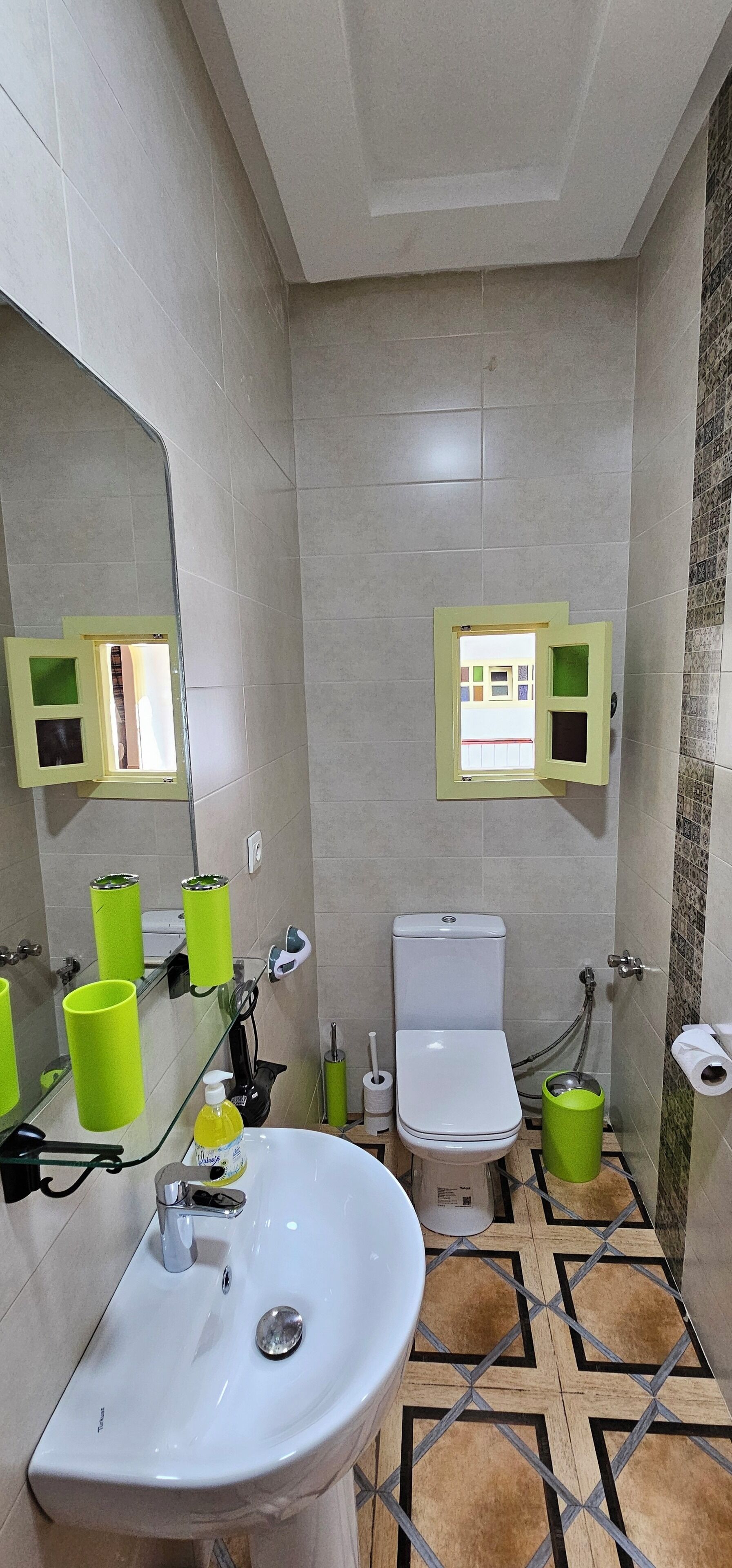 A bathroom with a white sink, toilet, and bidet. There are green trash bins and a mirror with a yellow frame. The floor has a geometric tile pattern.