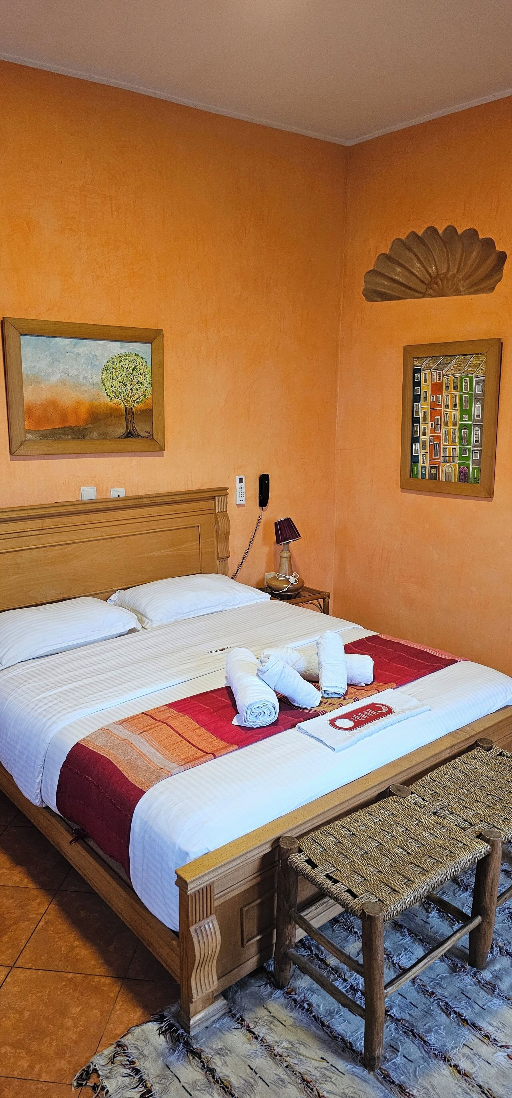 Een netjes opgemaakt bed met witte lakens, een rood-oranje gestreept dekbed en opgerolde handdoeken op een houten hoofdbord. Er staat een houten nachtkastje met een lamp en een telefoon.