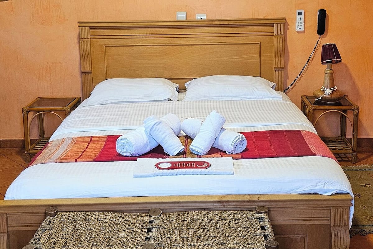 Una cama bien tendida con toallas, un cabecero de madera, un cuadro de un árbol y un teléfono en la mesita de noche.