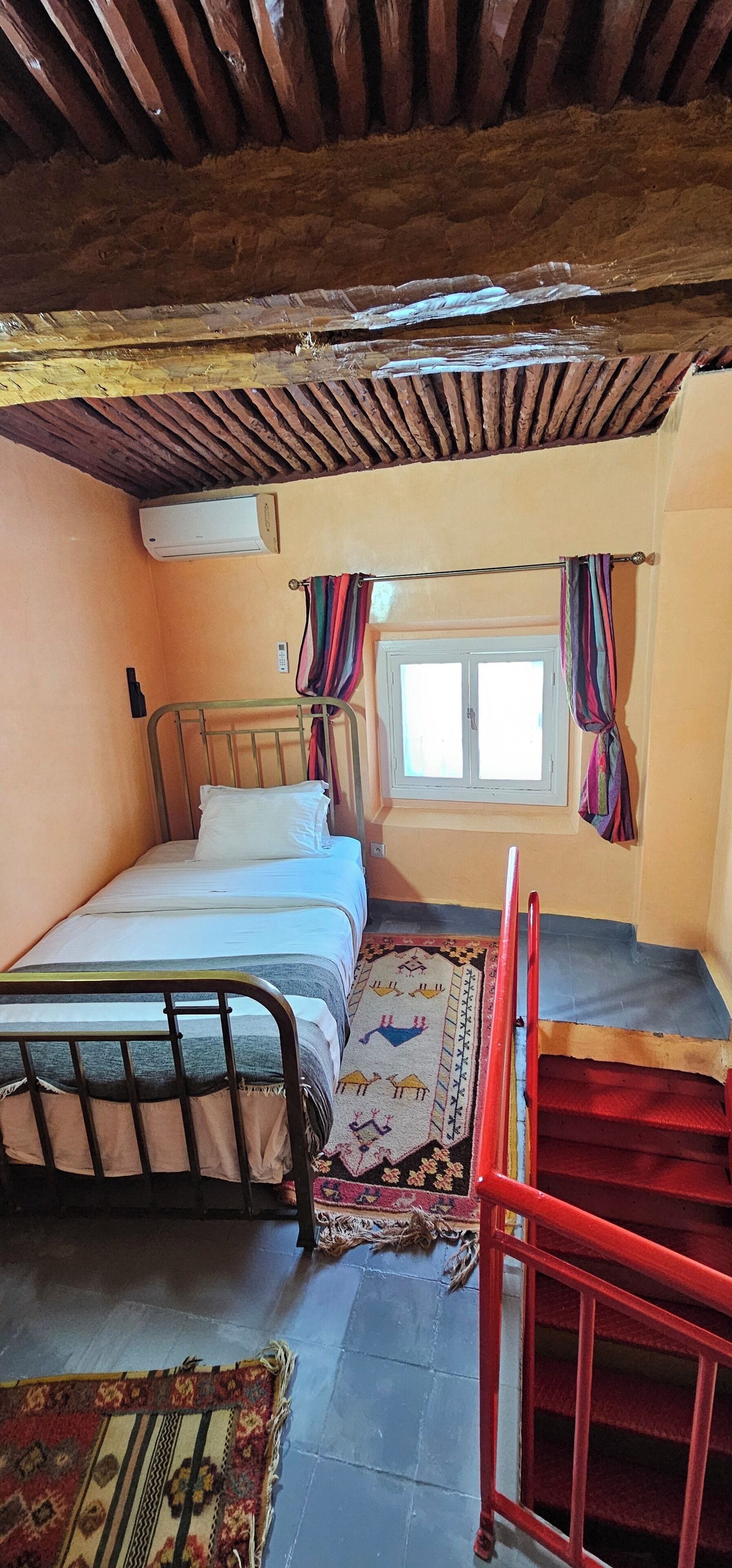 Una habitación pequeña y acogedora con una cama individual, una ventana con cortinas, una silla roja y una alfombra estampada.