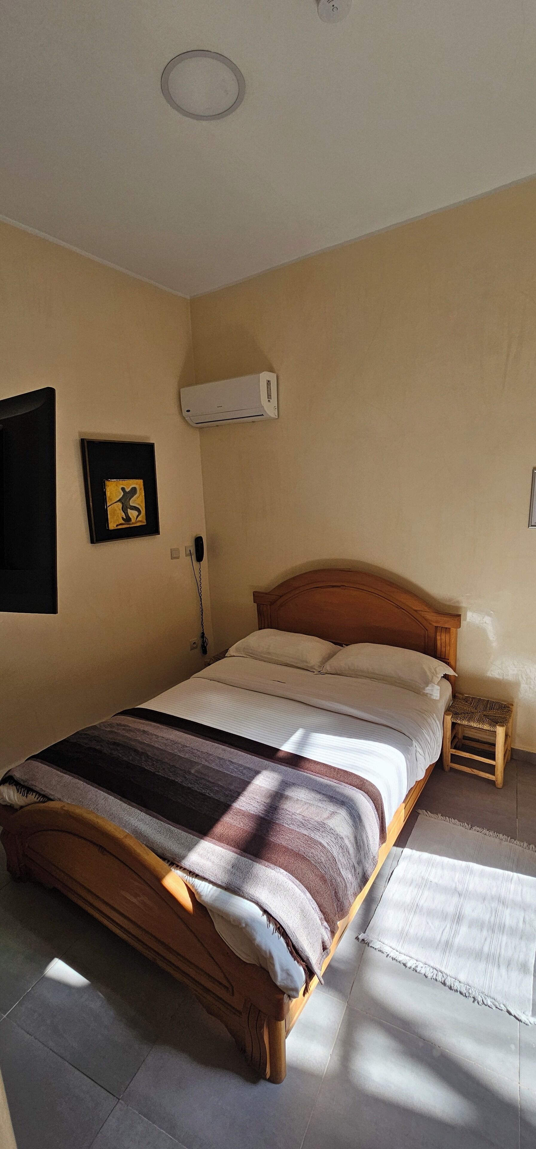 A bedroom with a wooden bed, a wall-mounted air conditioner, a framed picture, and a bedside table.