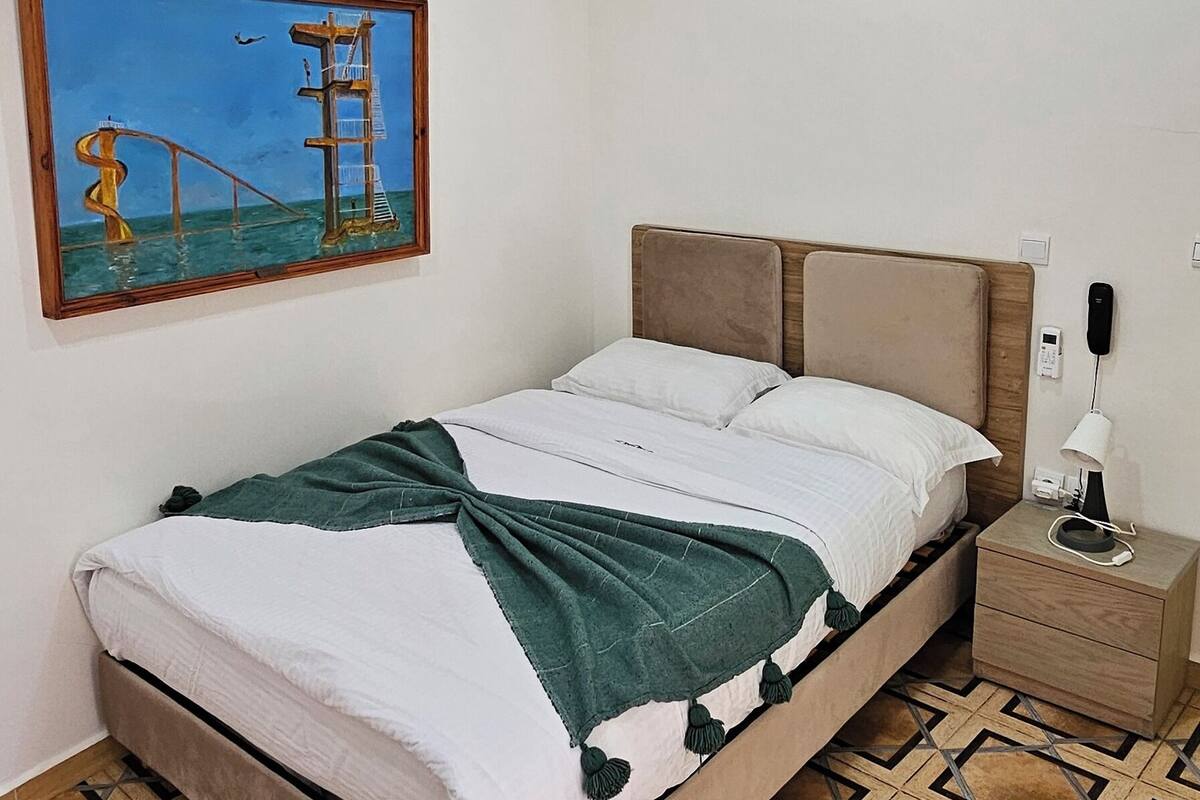 Un dormitorio con cama, mesita de noche, unidad de aire acondicionado, un cuadro colgado en la pared y suelo estampado.