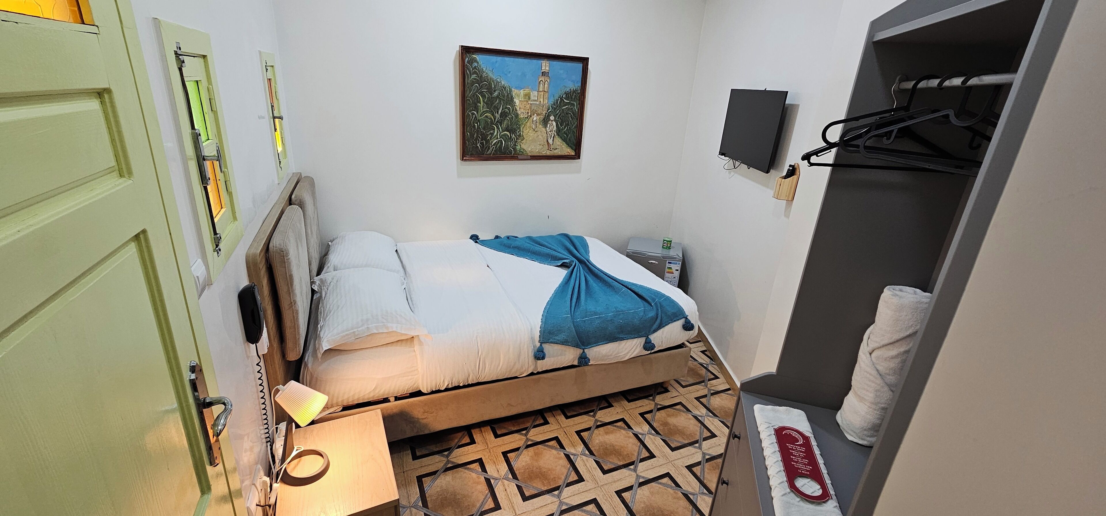 A small, neatly arranged bedroom with a bed, bedside table, lamp, and a painting on the wall.