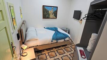 A small, neatly arranged bedroom with a bed, bedside table, lamp, and a painting on the wall.