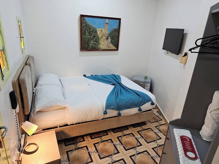 A small, neatly arranged bedroom with a bed, bedside table, lamp, and a painting on the wall.