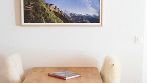 Une pièce avec une table en bois, deux chaises blanches dotées d’assises moelleuses, et un tableau encadré représentant un paysage de montagne accroché au mur.