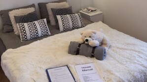 Une chambre à coucher avec un lit, des oreillers, un ours en peluche, une lampe, une étagère fixée au mur, un tableau encadré et un luminaire fixé au mur.
