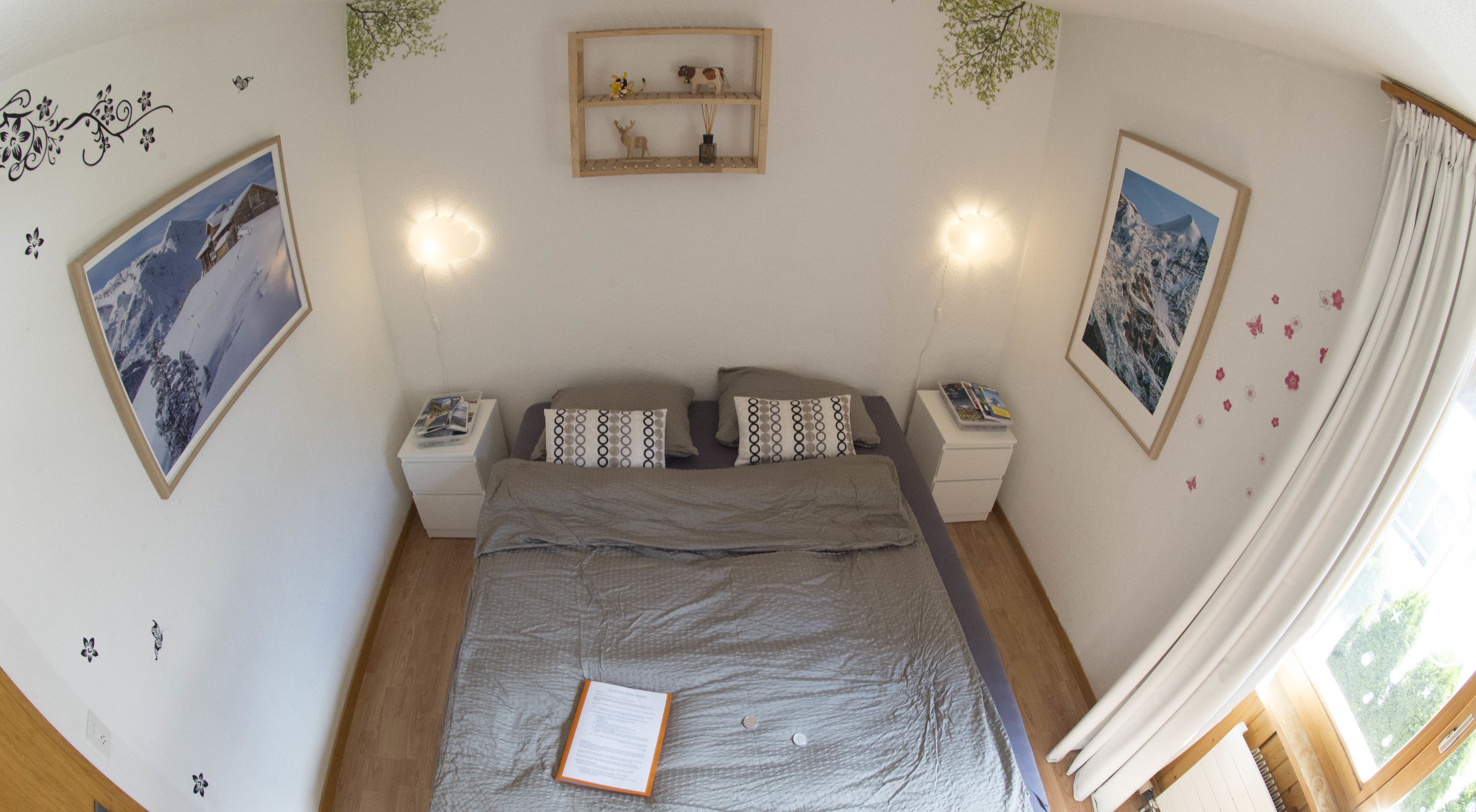 Un dormitorio con cama, dos mesitas de noche, un buró, un estante de pared con adornos y dos cuadros enmarcados.