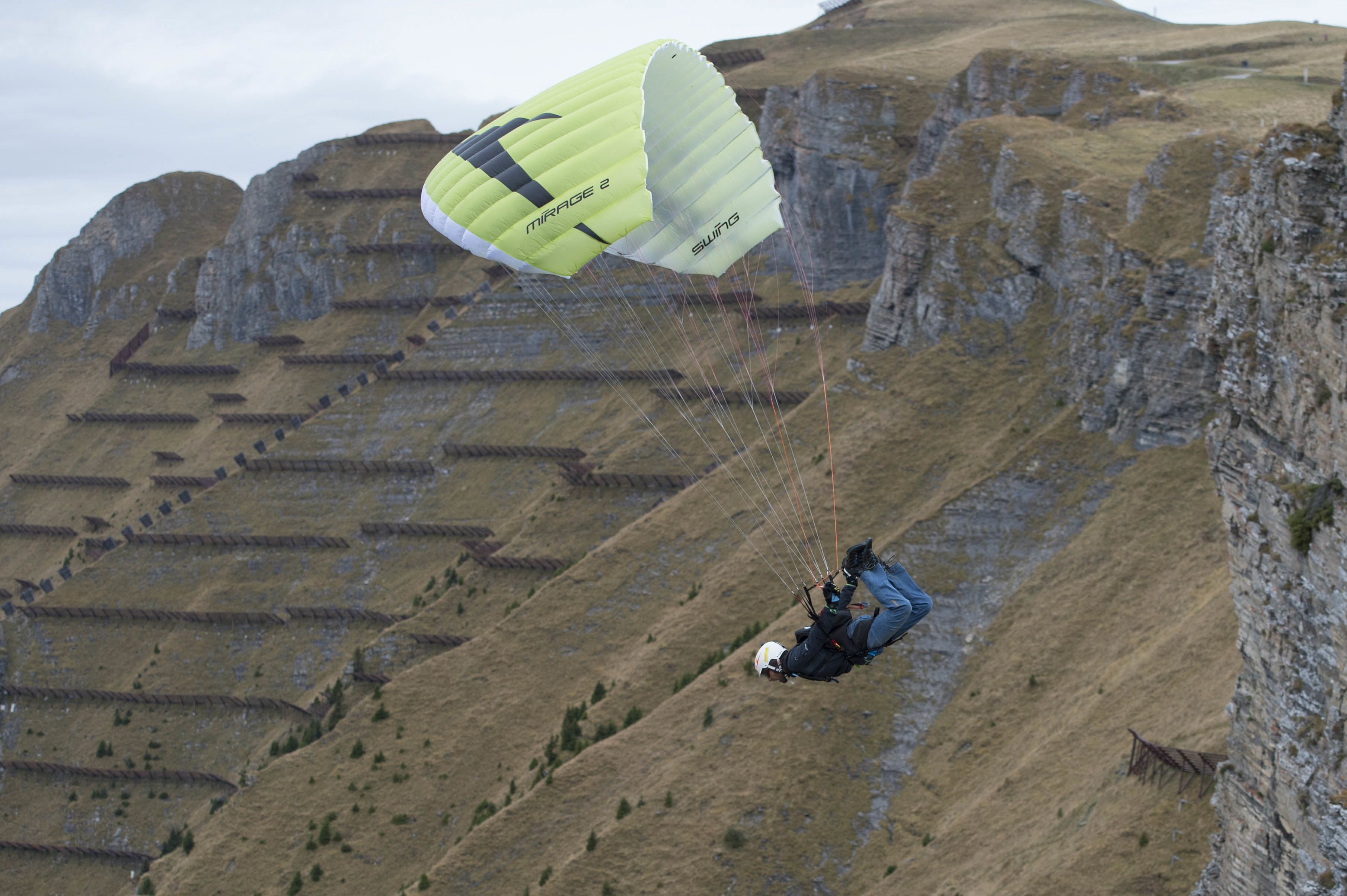 Une personne pratiquant le parapente avec un parachute vert et blanc au-dessus d’un paysage montagneux.