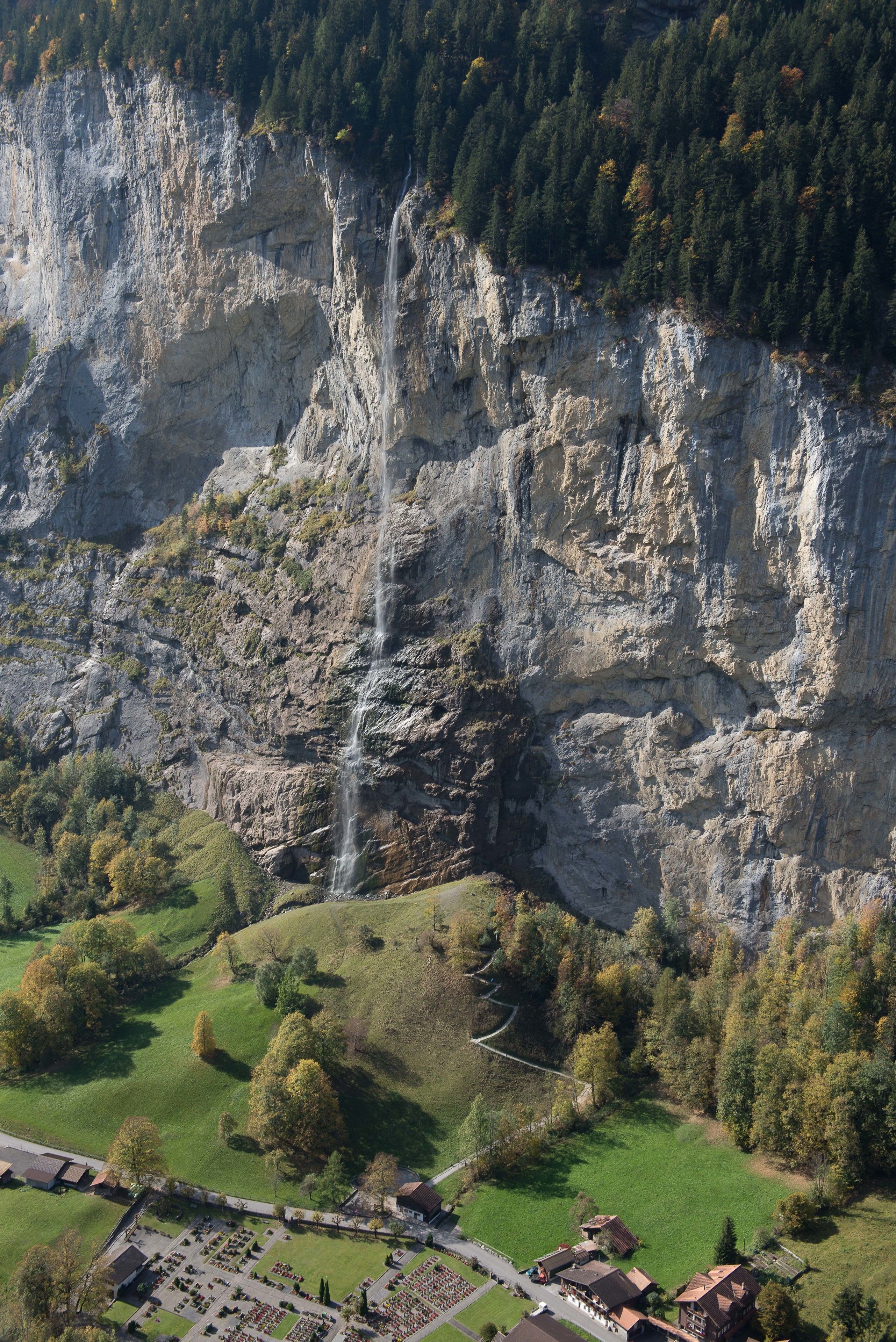 A mountainous landscape with a waterfall, green fields, and a small village with buildings and a parking area.