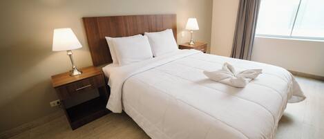 A hotel room with a large bed, two bedside tables with lamps, a wooden headboard, and a window with curtains.