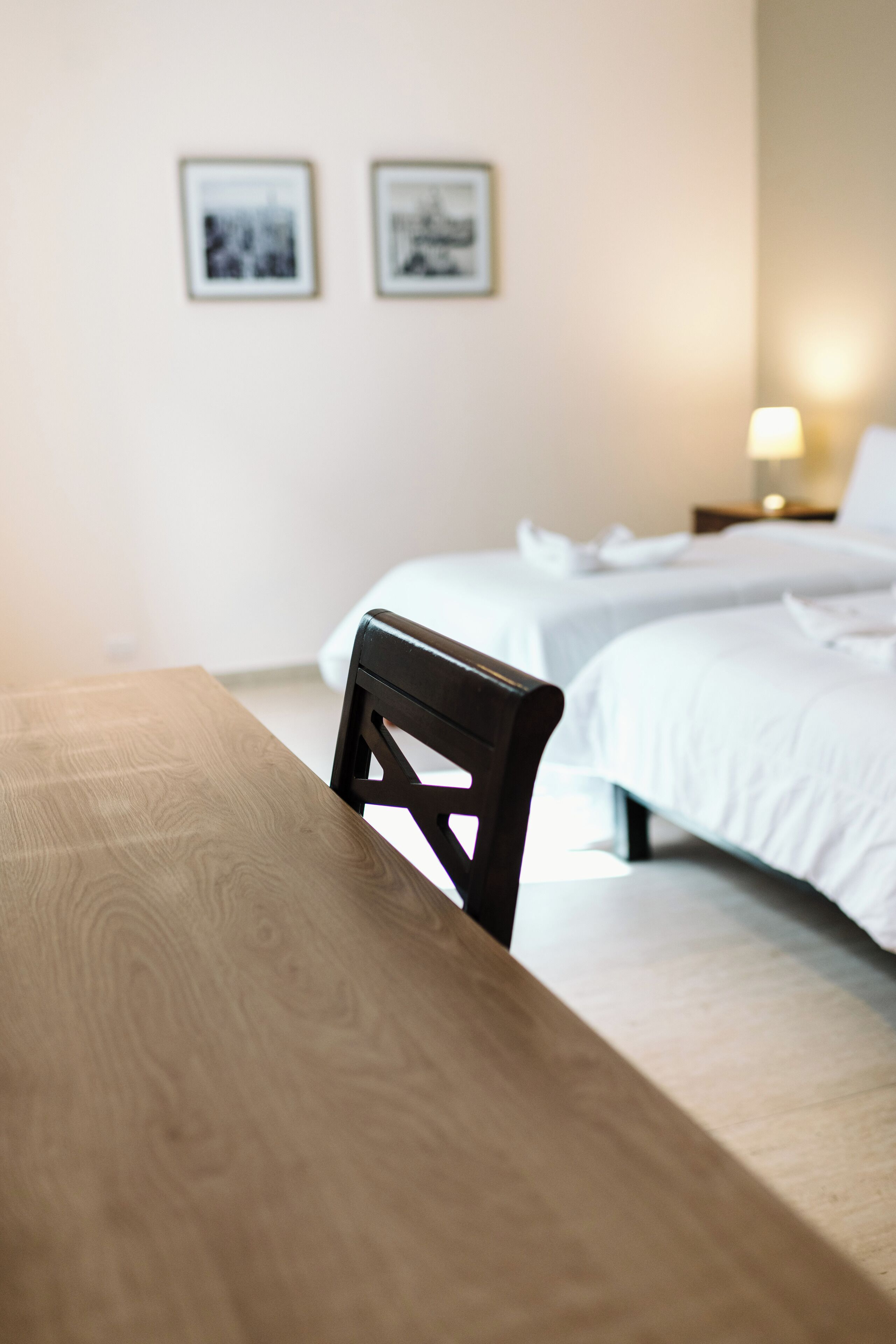 A hotel room with a wooden table and a black chair in the foreground, two beds with white bedding in the background, and two framed pictures on the wall.