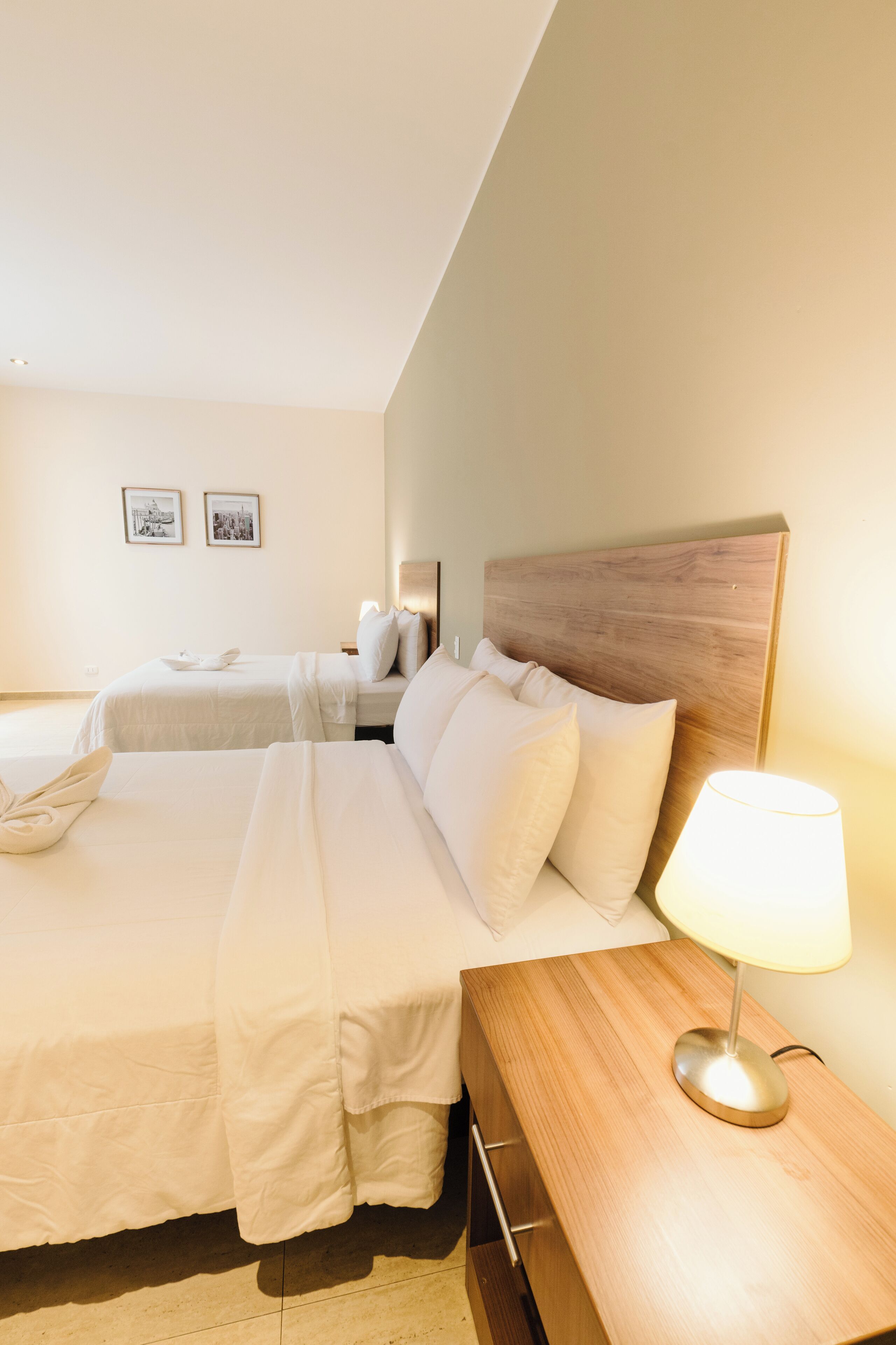 Una habitación de hotel con dos camas, cabecera de madera, lámpara de noche y una mesita.