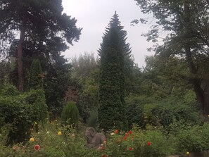 A garden with a variety of trees, a cross-shaped stone, and a ram statue amidst flowering plants.