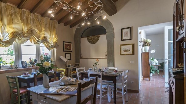 A dining area with a rustic wooden ceiling, a brick floor, and a large window with yellow curtains. There are several tables and chairs, and a vase with flowers on one table. - B&B La Chiusa Delle Monache (Lucca)