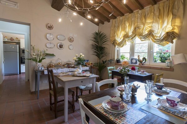 Un comedor acogedor con una mesa de madera dispuesta para una comida, rodeada de sillas. Se ve una cocina al fondo, y hay platos decorativos en la pared.