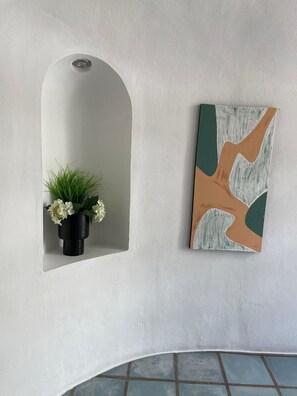 Una habitación con pared blanca con textura, un florero negro con flores blancas y un cuadro abstracto geométrico.