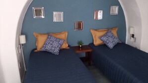 Quarto com duas camas, cada uma com colcha azul e almofadas estampadas, uma pequena mesa de madeira com uma planta e uma luminária na parede.