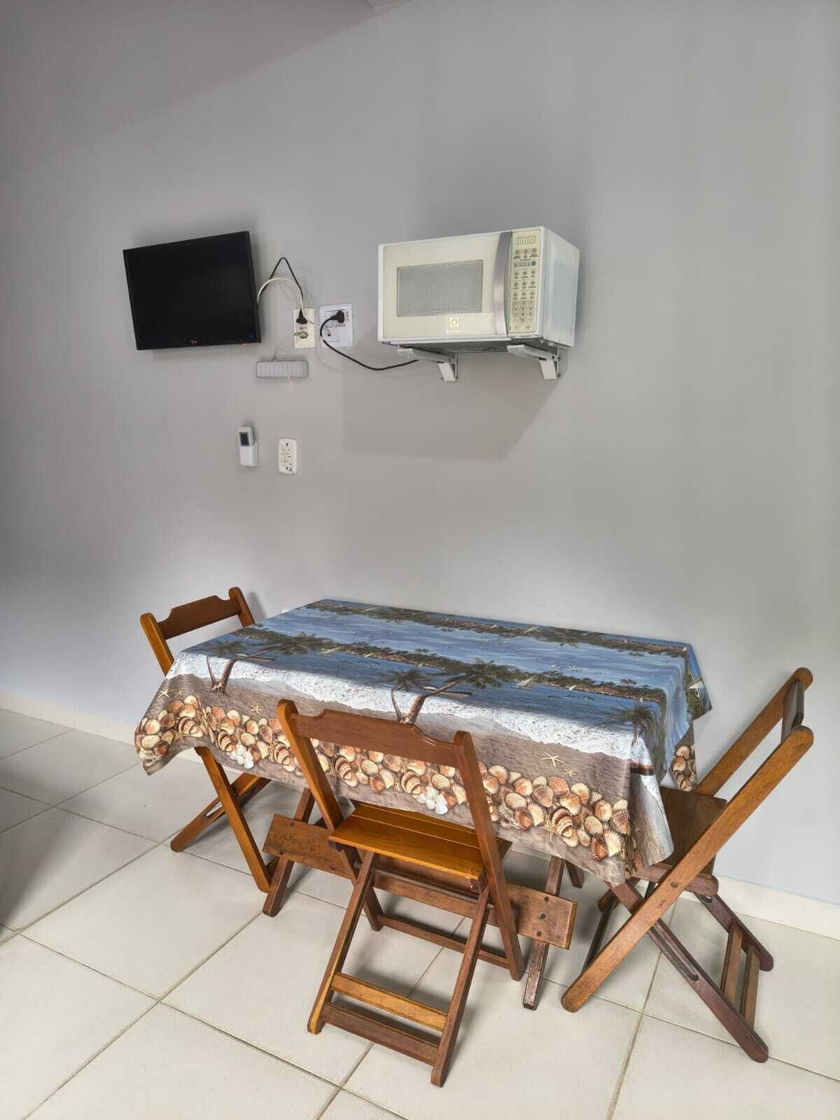 A dining table with a beach-themed tablecloth, wooden chairs, a television, and a microwave on a wall.