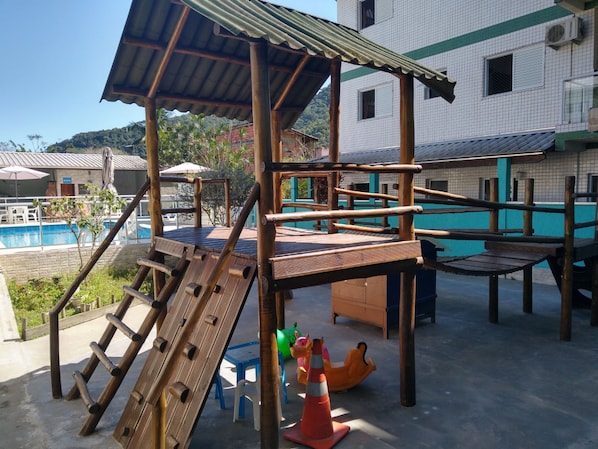 Una estructura de juegos de madera con toboganes y elementos para escalar, una piscina y un edificio con fachada de azulejos.