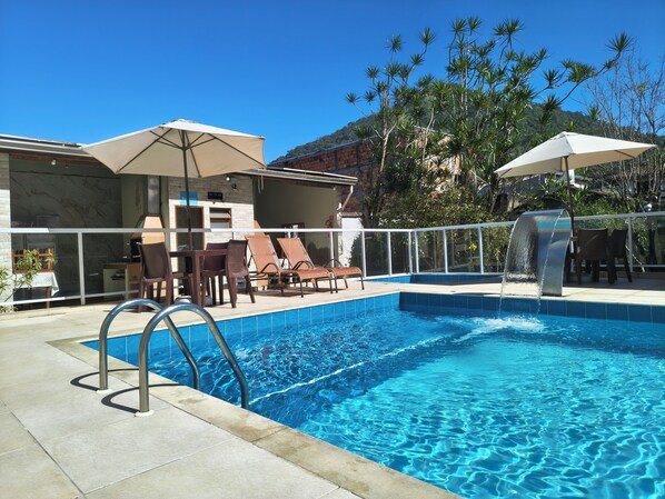 A swimming pool with a clear blue water surface, surrounded by lounge chairs and umbrellas, with a view of a mountain in the background.