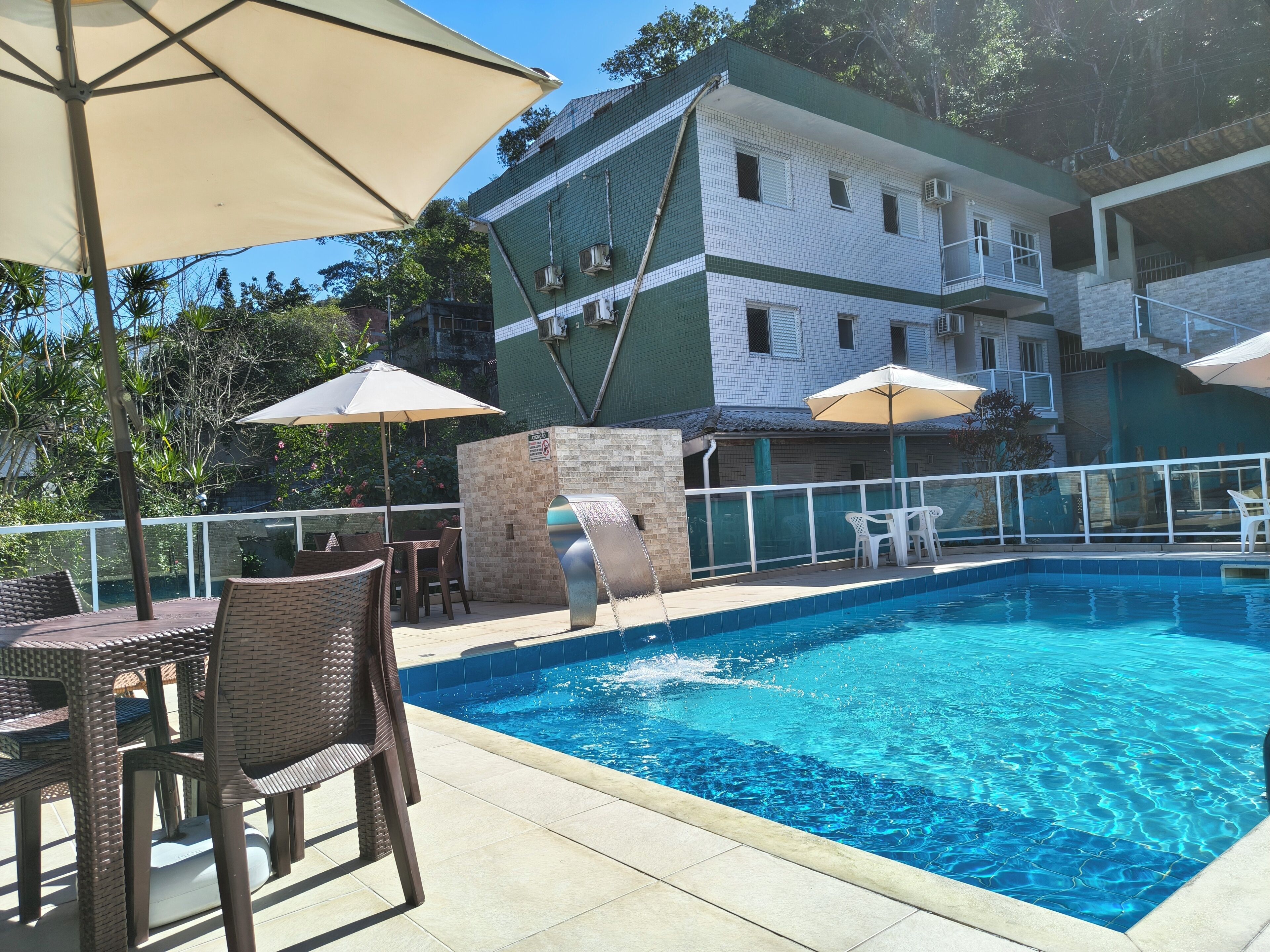 A swimming pool with a tiled edge, surrounded by outdoor furniture and umbrellas, with a building in the background.