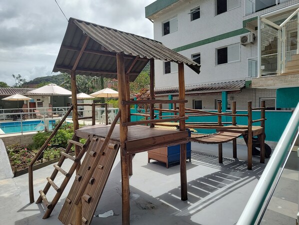 A wooden play structure with a roof, a pool, and a building with a tiled exterior in the background.